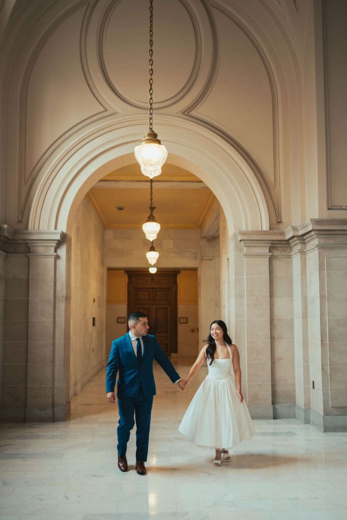 A newly married couple walks hand in hand beneath the grand arches of San Francisco City Hall during an intimate elopement.