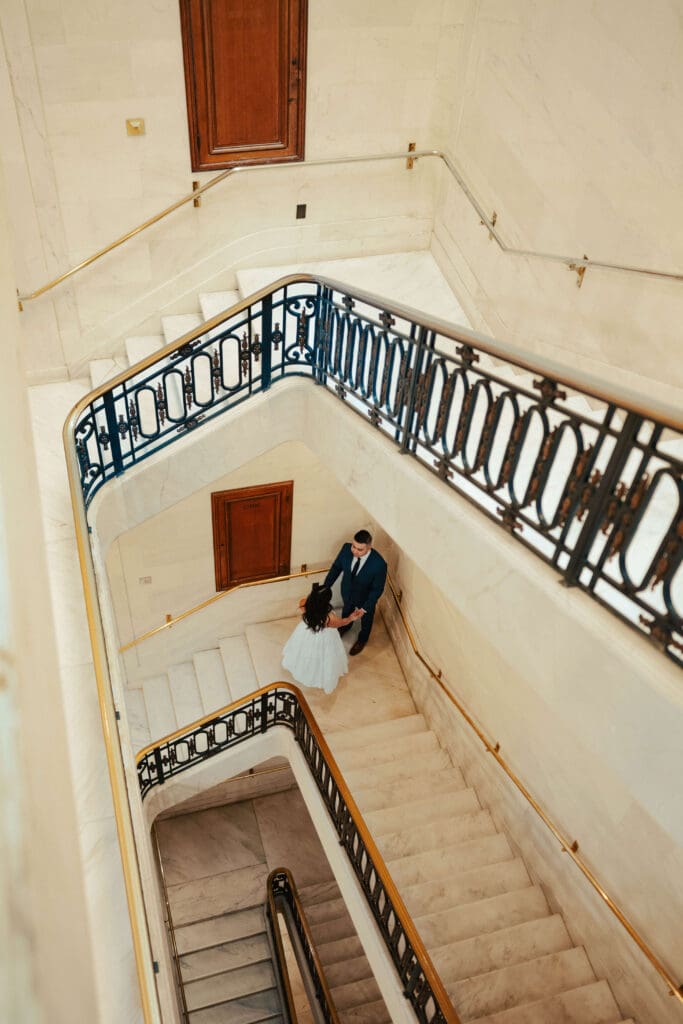 A cinematic overhead view of a couple holding hands on the iconic grand staircase at San Francisco City Hall.