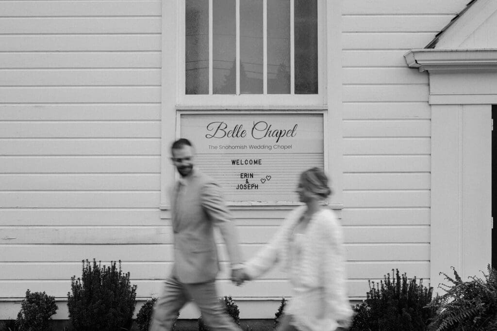 The couple walks hand in hand past a Belle Chapel sign, captured with motion blur in black and white.