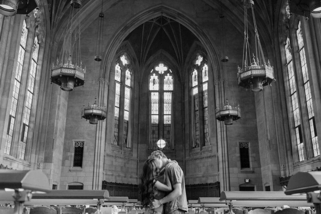 Black and white photo of a couple kissing between reading tables under vaulted ceilings and chandeliers