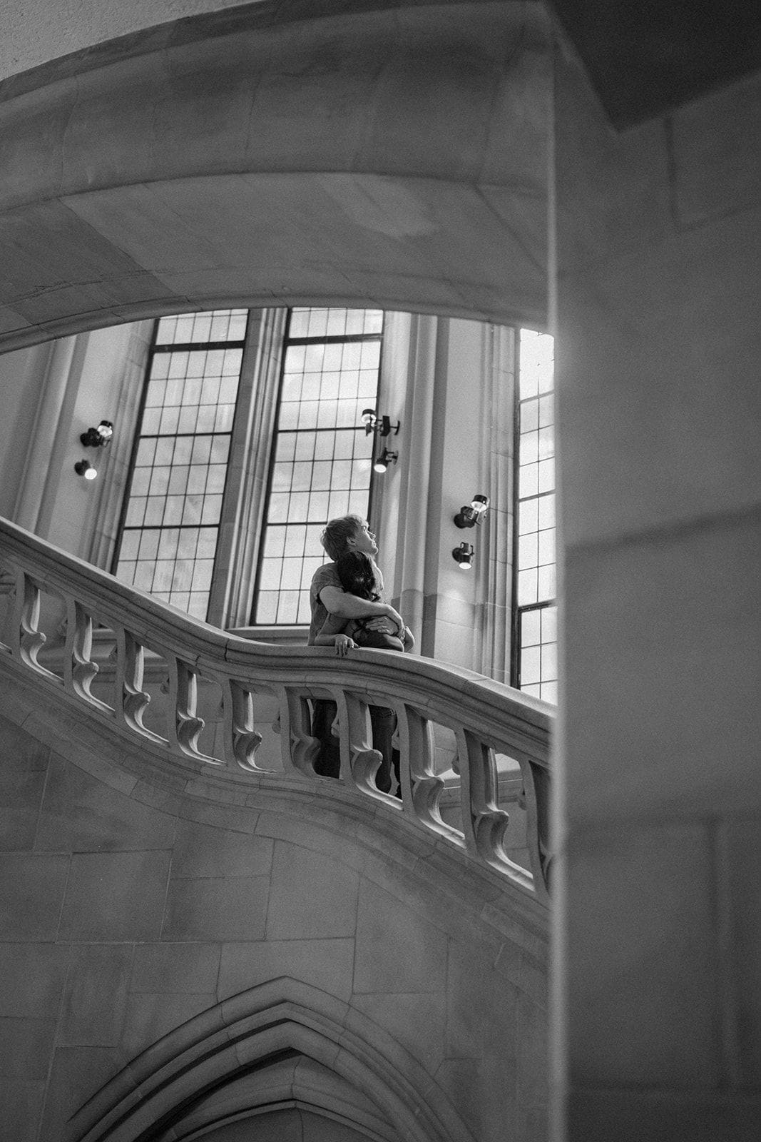 Black and white photo of a couple embracing on a curved stone staircase, framed by a gothic arch and tall windows at Suzzallo Library in Seattle