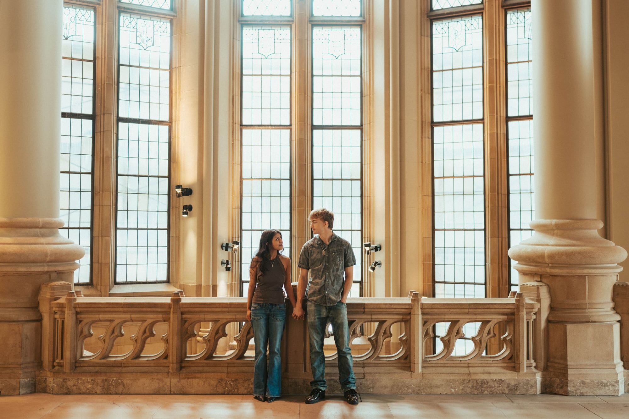 Couple leaning against a carved stone railing and holding hands under tall arched windows in Seattle