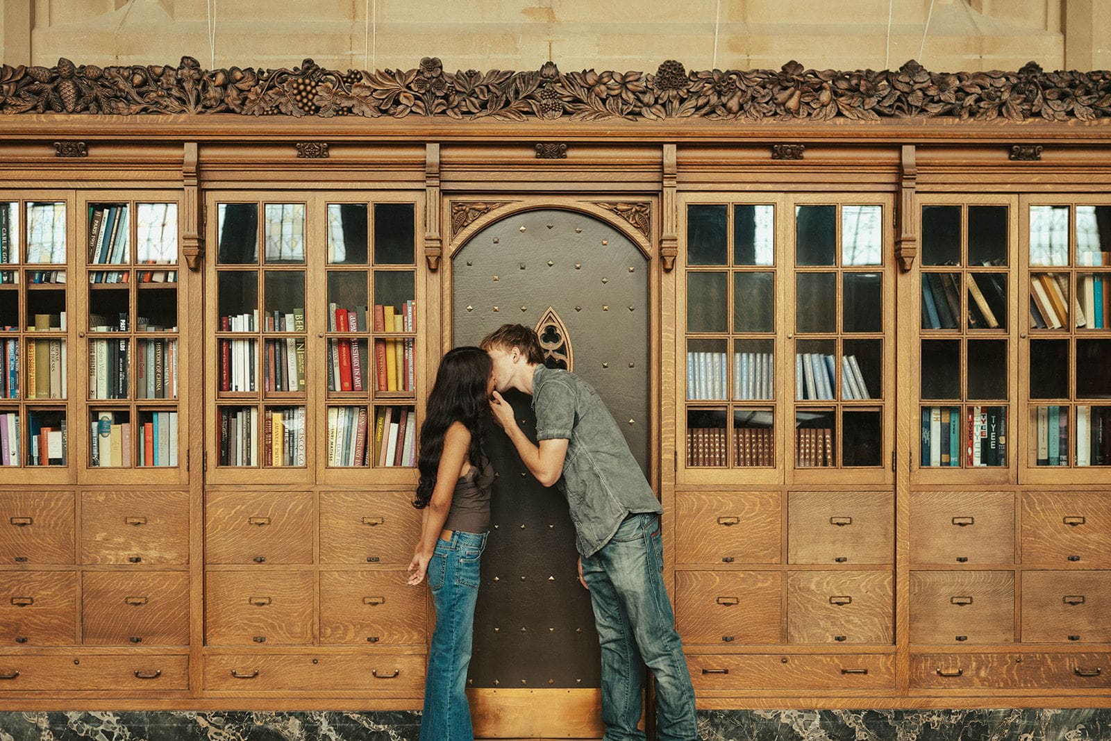 Couple kissing in front of a studded gothic door framed by carved oak cabinets and floral molding at Suzzallo Library