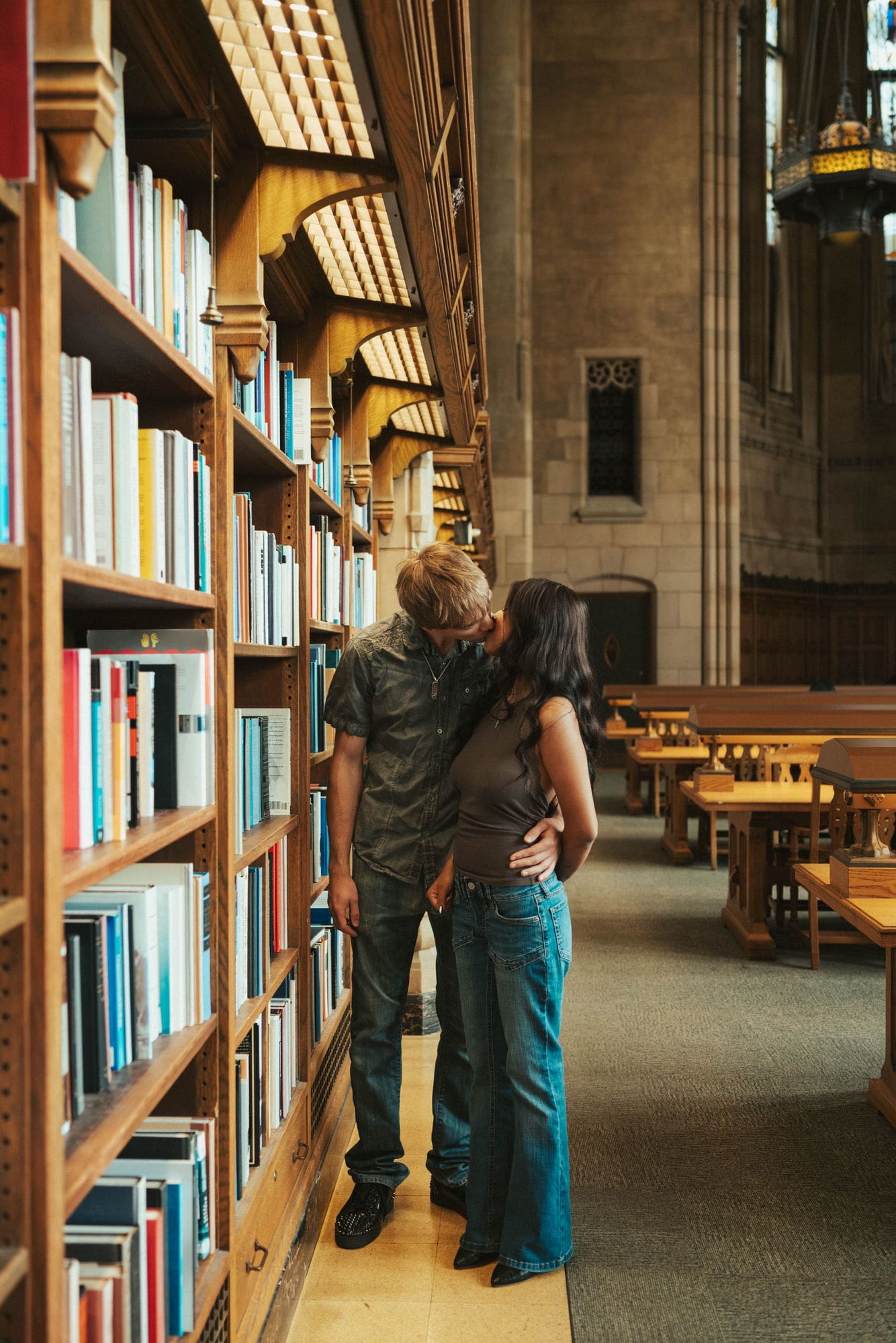 Couple kissing in a narrow aisle of library book stacks with warm light on the wooden shelves