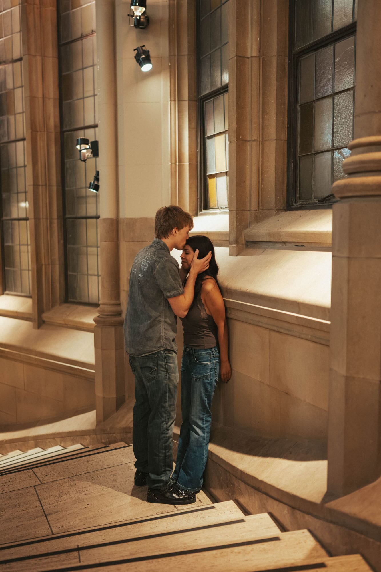 Man cupping his partner's face and kissing her forehead in a sunlit stone corridor at Suzzallo Library
