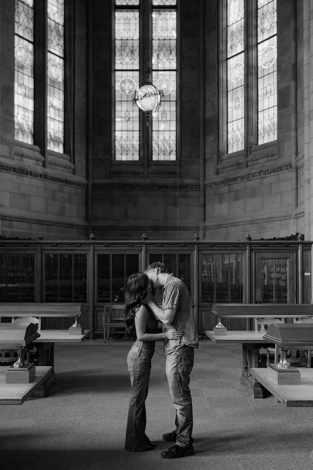 Black and white photo of a couple in a quiet embrace beneath towering stained glass windows in the Suzzallo Library reading room