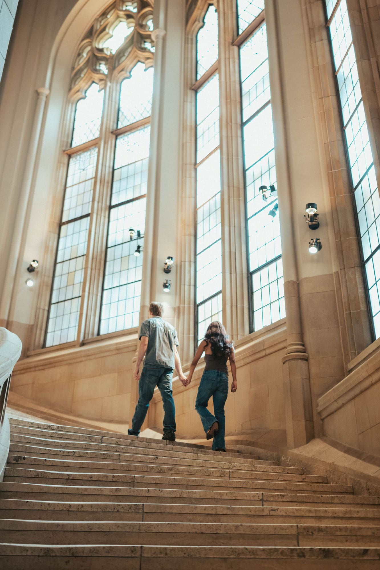 Couple holding hands as they climb a wide stone staircase beneath towering gothic windows at Suzzallo Library in Seattle