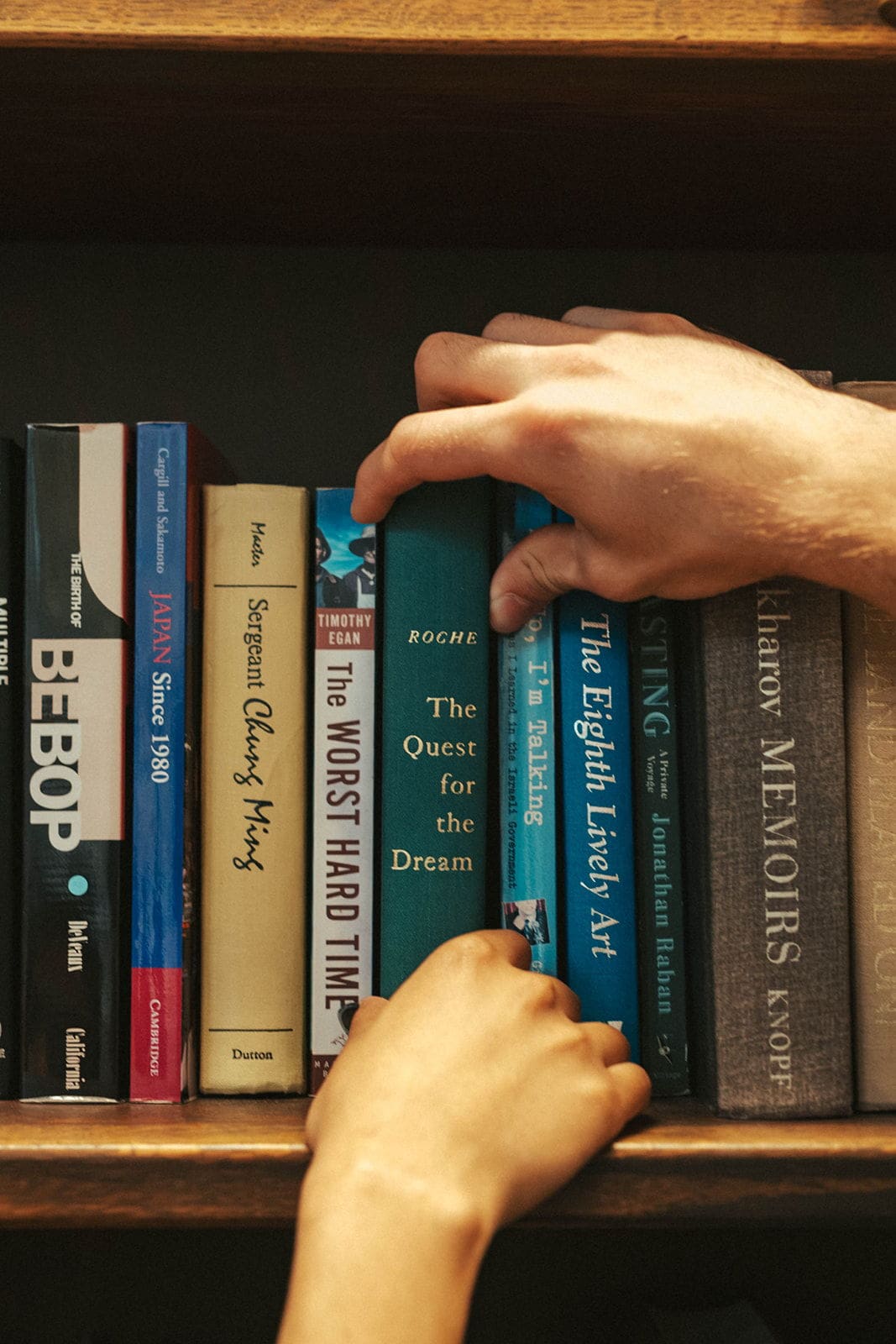 Two hands reaching for the same book on a wooden library shelf, fingers overlapping on the spine of a green hardcover