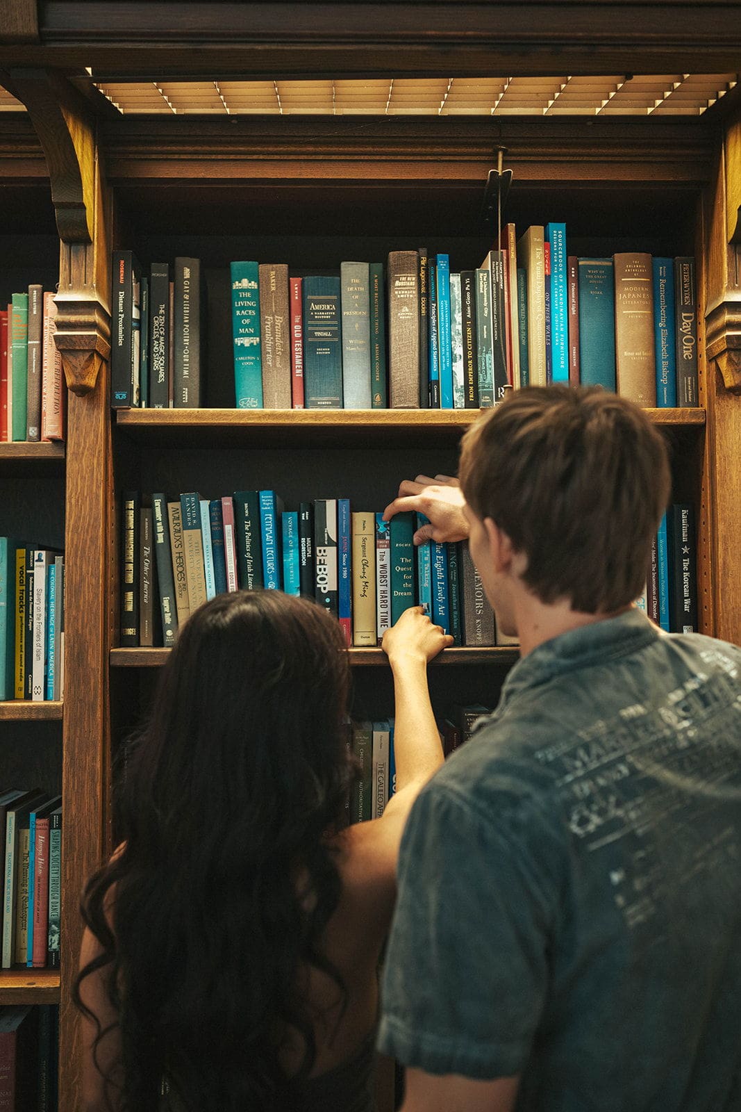 Couple seen from behind reaching for the same book on a tall oak shelf inside Suzzallo Library in Seattle